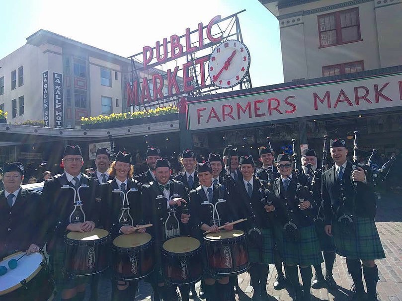 members from the Keith Highlanders Pipe Band pose in front of the famous Public Market Center clock & sign at Pike Place Market
