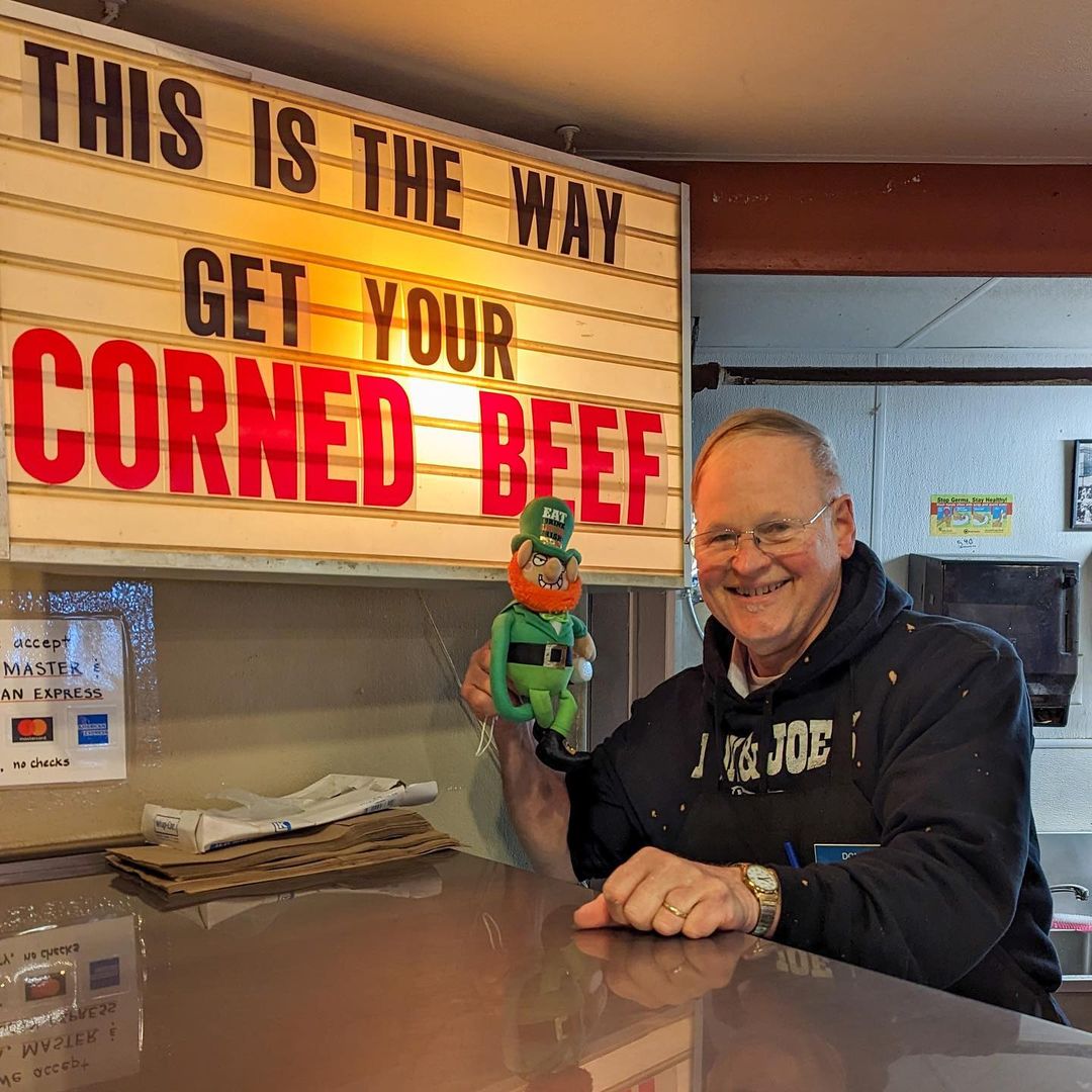 Owner don from don & joe's meats in Pike Place Market stands by a marquee that reads this is the way get your corned beef