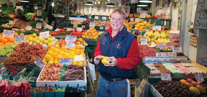 Mike Osborn, owner of Sosio's Fruit & Produce, stands proudly at the renowned Pike Place Market produce stand. Clutching a vibrant piece of fresh fruit, he embodies the commitment to quality and tradition that has defined Sosio's over last 80 years. The bustling market atmosphere and colorful array of fruits provide a glimpse into the rich experience of this longtime establishment.